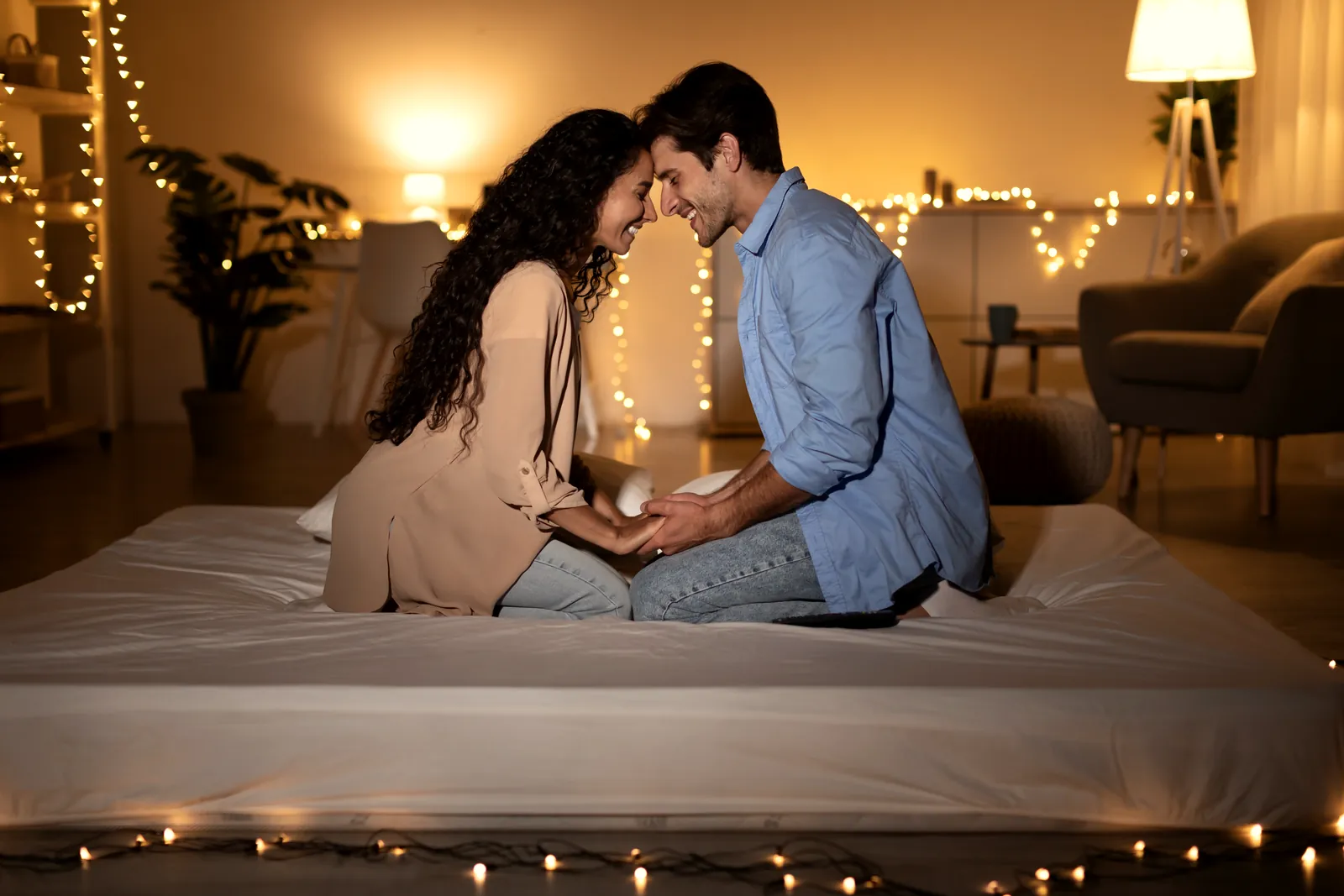 A couple sitting on a bed, foreheads touching, in a warmly lit room.